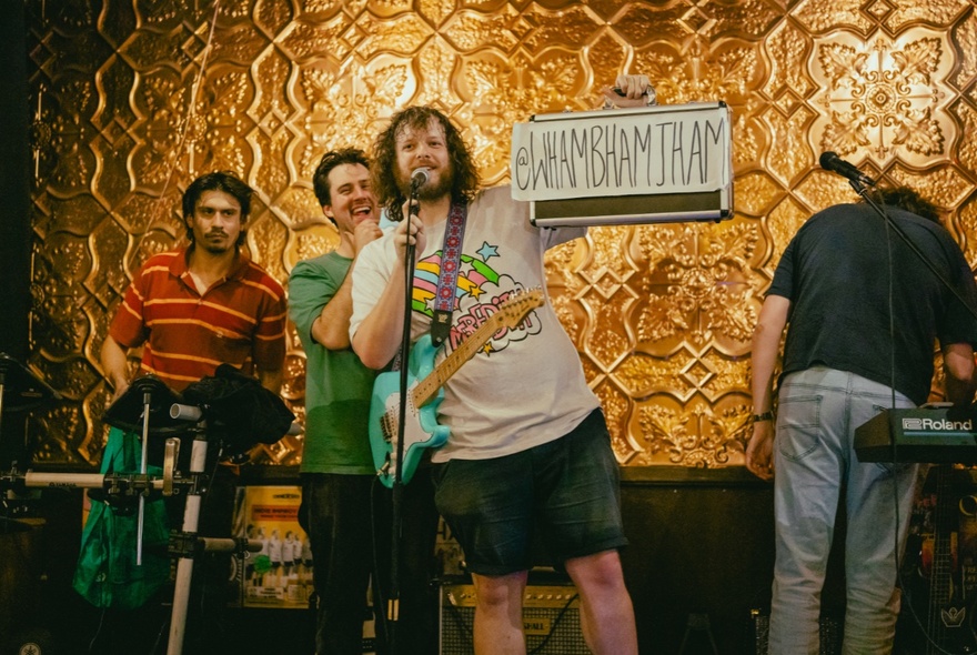 Four people on a stage, two of them with instruments, and one of them holding up a hand written sign that reads WHAMBHAMJHAM.