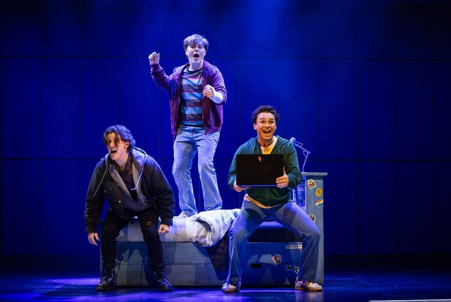 Three actors on a theatre stage, one of them standing and bouncing on a bed.