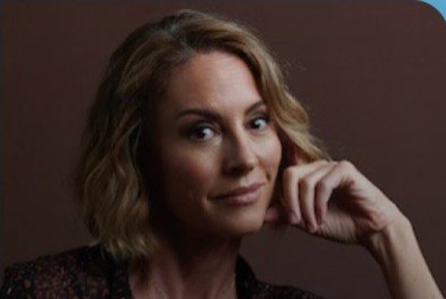 New York Times bestselling author, Sarah Wilson, smiling as she rests her face on her hand; brown background.