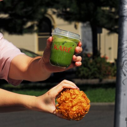 Hand appearing around a corner, one holding a matcha drink and another holding a bun. 