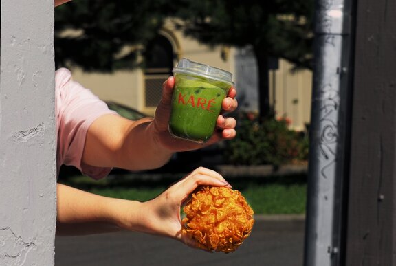 Hand appearing around a corner, one holding a matcha drink and another holding a bun. 