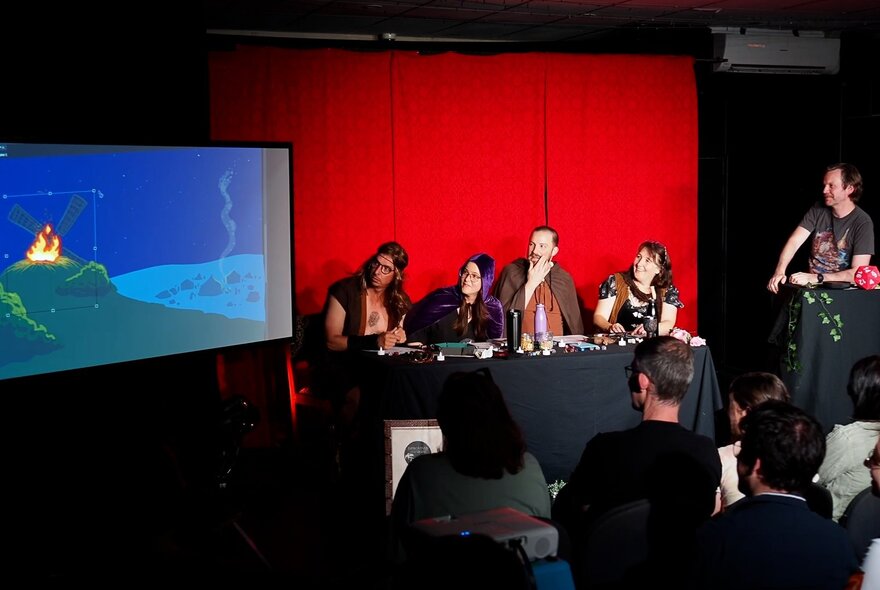 Four performers seated at a table on a stage behind a table, with a person guiding them through a Dungeons and Dragons style improvised adventure, and a large screen on the stage featuring a projected image. 