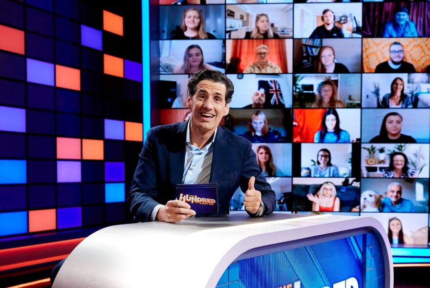 Andy Lee seated behind TV desk with cards and thumbs up, video screens in the background.