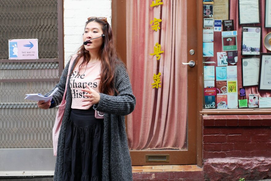 A woman speaking through a mouth microphone, holding paper and gesticulating.
