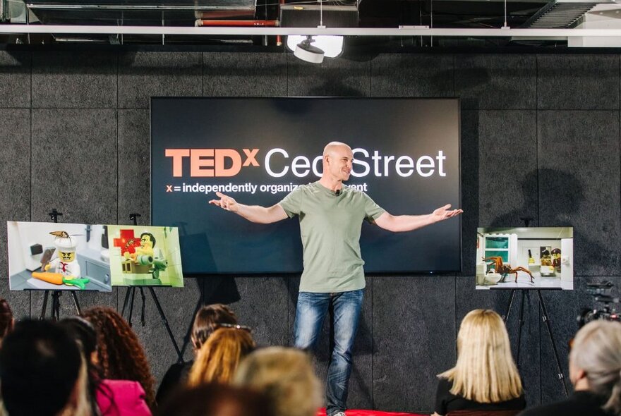 A man standing in front of an audience with arms outstretched, with photos of Lego diaramas and Australian wildlife on either side.