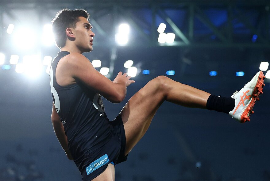 A Carlton footballer kicks his leg high during a match under lights. 