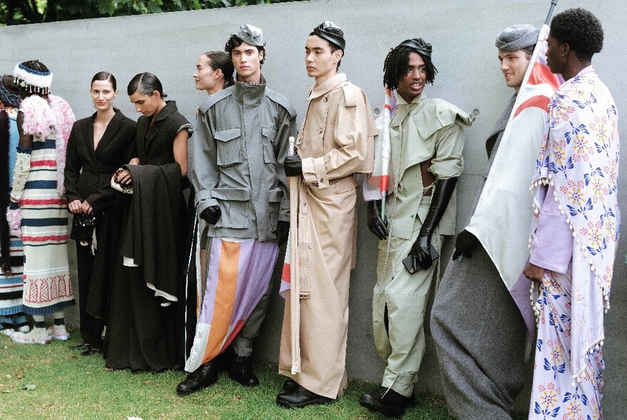 Models back stage at a fashion show leaning against a wall in elaborate attire.