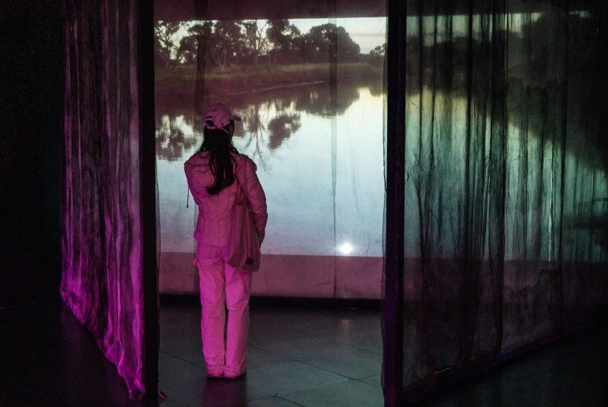 A woman standing looking at a projection of water with sheer curtains either side, a pink light cast on her.