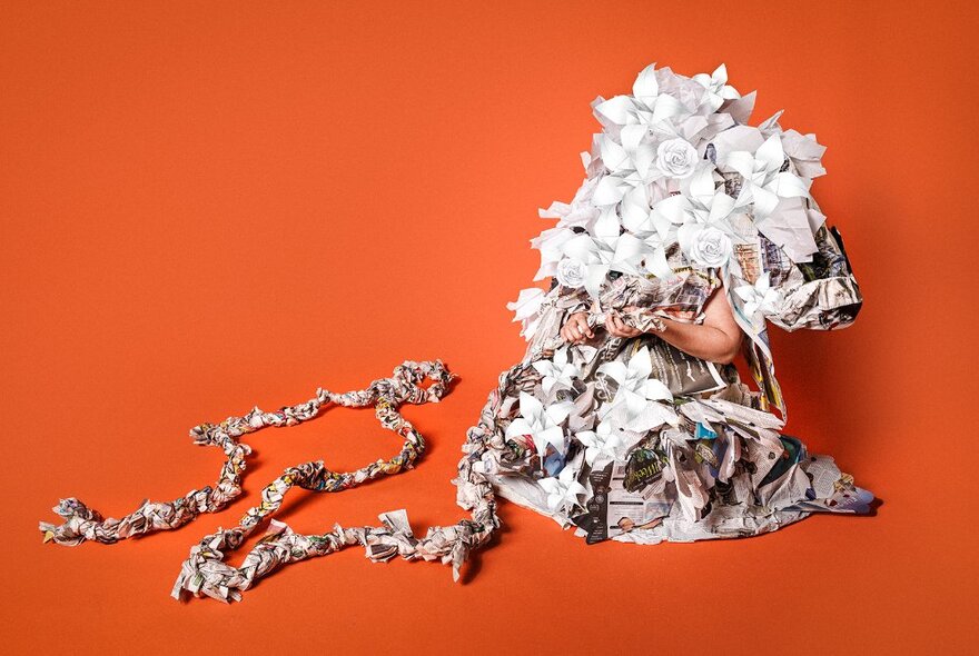 A person seated on the ground completely covered in an outfit made from pieces of folded newspaper and white paper in the form of flowers, only their forearms visible.