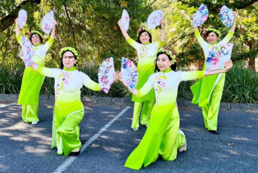 Children in traditional Chinese green and white dresses performing outside in dappled shade, waving fans.