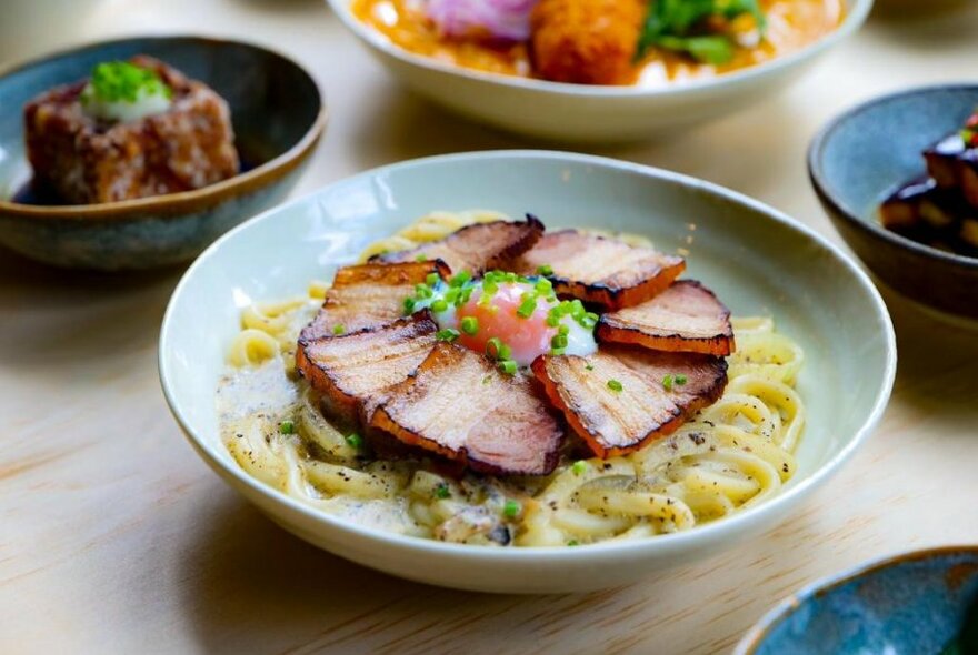 Meat and egg on noodles in bowl, surrounded by several other dishes.