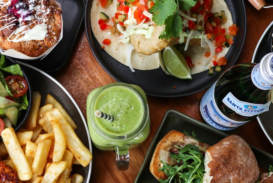 Bird's-eye view of green smoothie, chips, salad, pizza wrap and open sandwich on wooden table.