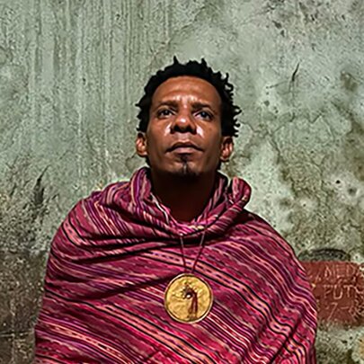A Timor-Leste man wearing traditional clothing and a circular gold pendant in front of a rough concrete wall.