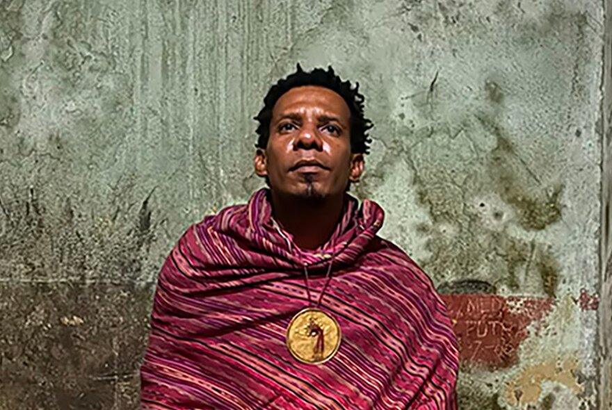 A Timor-Leste man wearing traditional clothing and a circular gold pendant in front of a rough concrete wall.