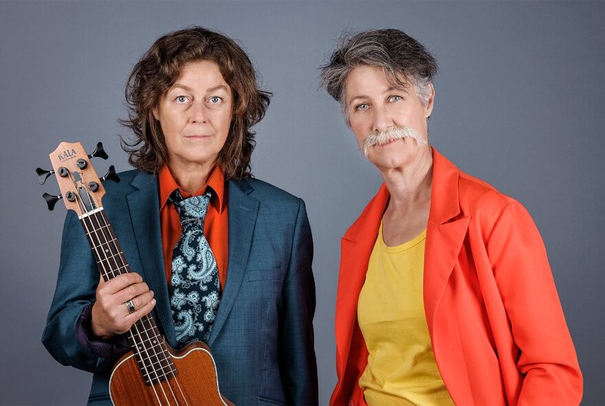 Two performers wearing suit jackets, one holding a guitar, the other wearing a fake moustache.