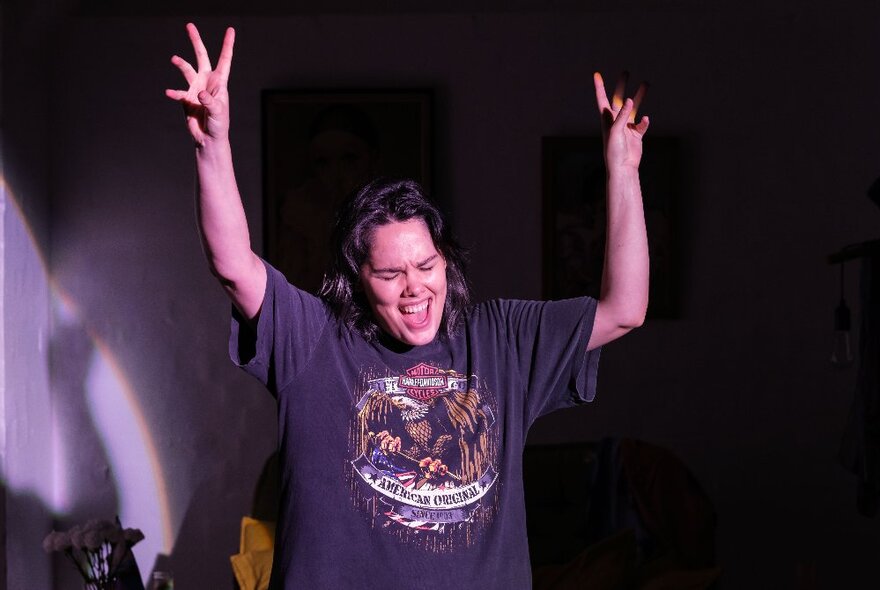 Comedian, Jenna Suffern, on stage in a black t-shirt, arms up in the air and smiling.