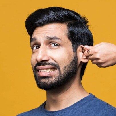 Comedian Kanan Gill, with short dark hair and a cropped beard, grimacing as a hand twists his ear.