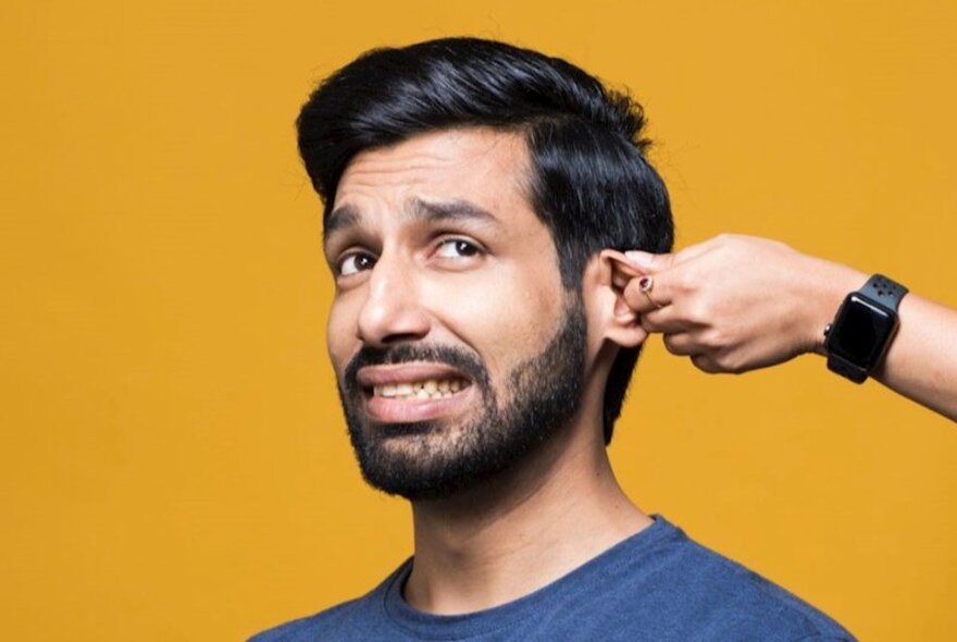 Comedian Kanan Gill, with short dark hair and a cropped beard, grimacing as a hand twists his ear.