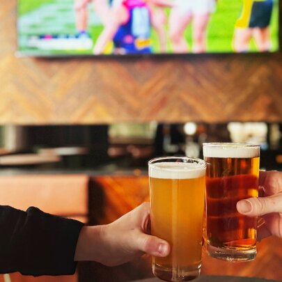Two hands clinking two glasses of beer together over a table, with a tv on the wall behind screening a game of footy in the background.