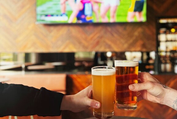 Two hands clinking two glasses of beer together over a table, with a tv on the wall behind screening a game of footy in the background.