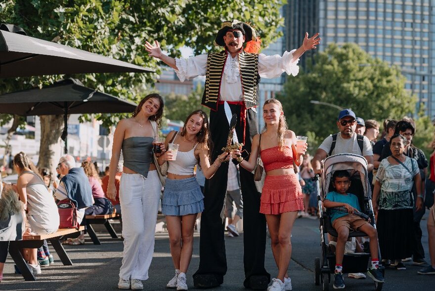 Three young women interacting with a roving entertainer, a man on stilts, in the outdoor environment of a summer night market, crowds walking around in the background.