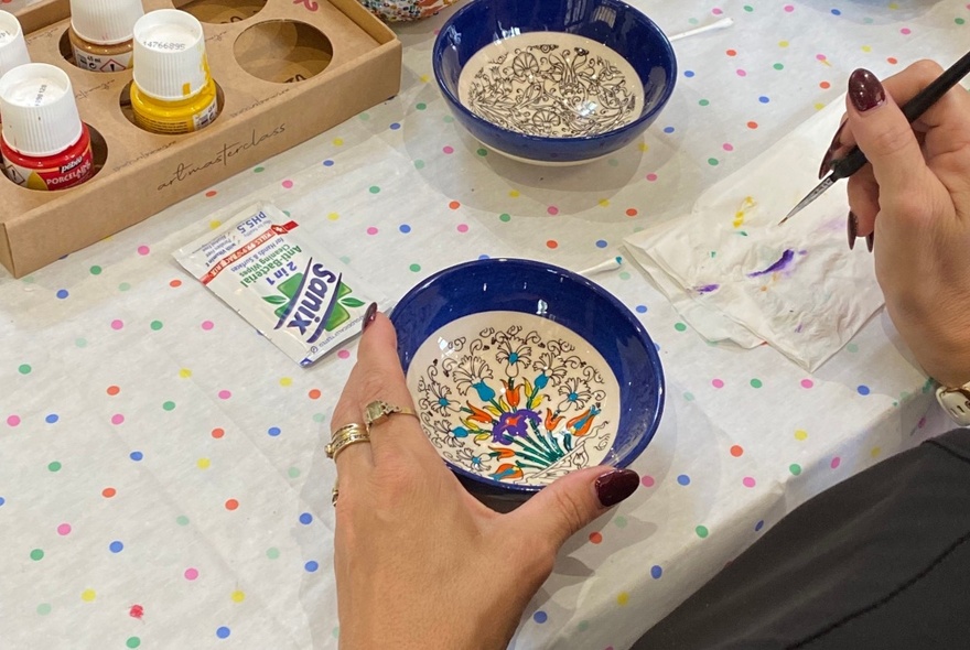 Hands holding a painted bowl in one hand, holding a brush with paint in the other, at a crafts table.