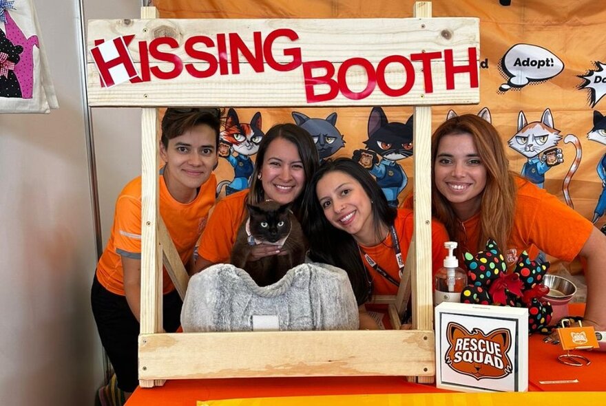 Four smiling people lined up and posing for a photo with a cat, underneath a sign that reads HISSING BOOTH.