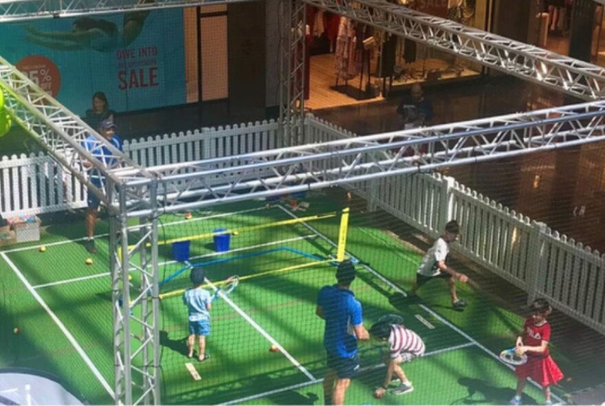 Kids and adults playing on a mini tennis court in a market setting.