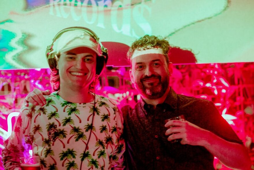 Two smiling people at an outdoor bar, one of them wearing headphones, the other holding a drink in his hand.