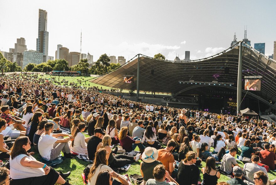 Crowd seated on grass during a daytime event at Sidney Myer Music Bowl, city buildings in the background.
