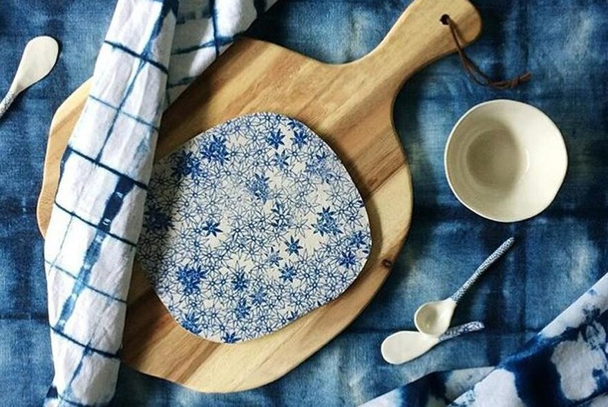 A small hand crafted ceramic bowl and spoons, displayed next to a wooden board, on a blue tie-dyed tablecloth at an arts and craft market stall.