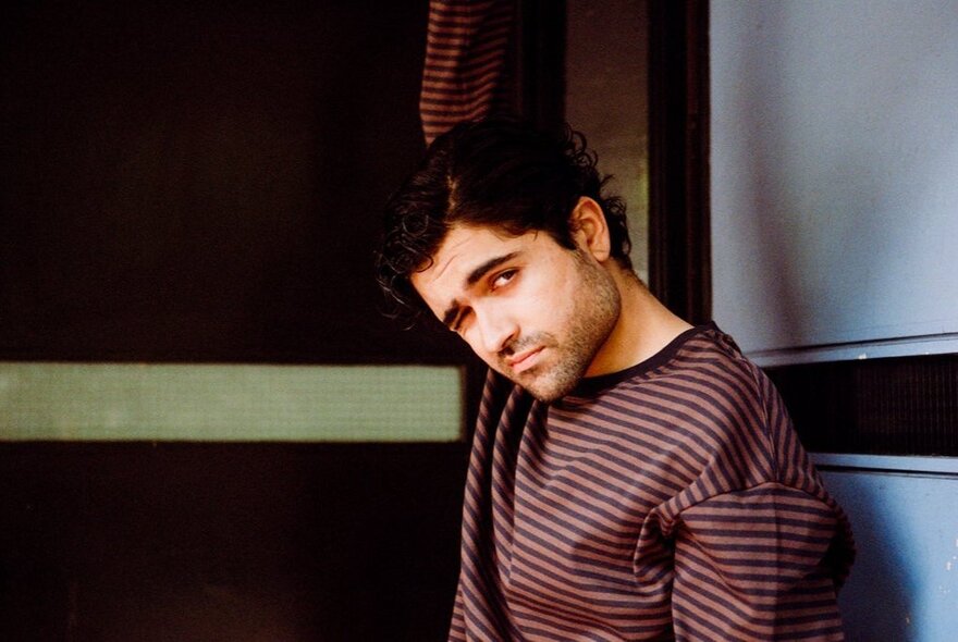 Singer Dhruv, wearing a striped top and looking moodily at the viewer while leaning against a wall.