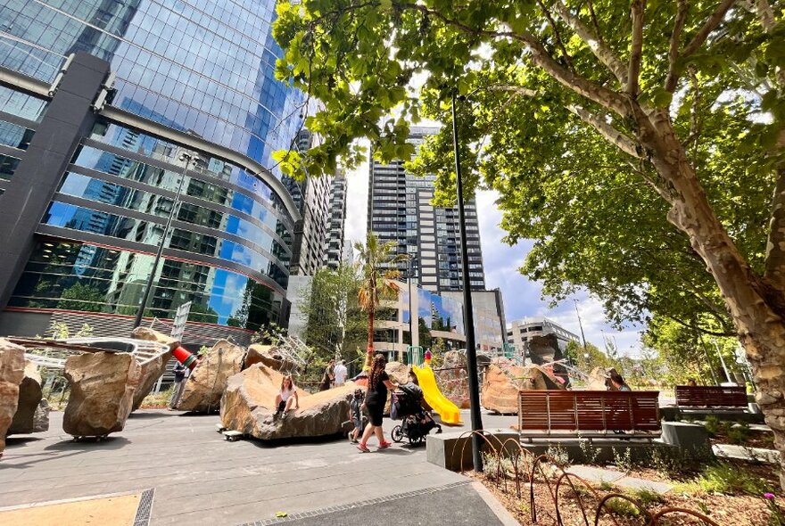 Woman pushing a pram through a city playground space that has large climbing rocks, ropes, swings, bench seats, with city skyscrapers in the background.