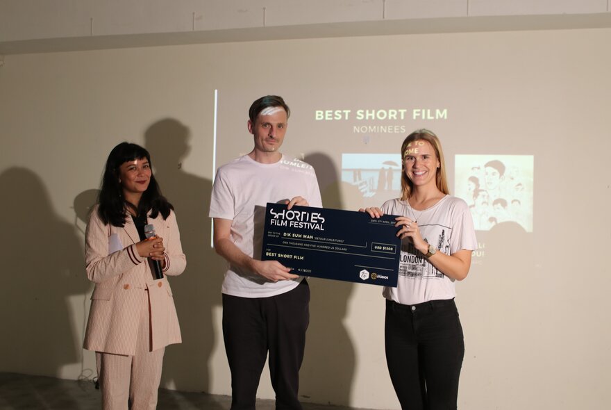 Three people in front of a projection screen, one accepting an award printed on large cardboard. 