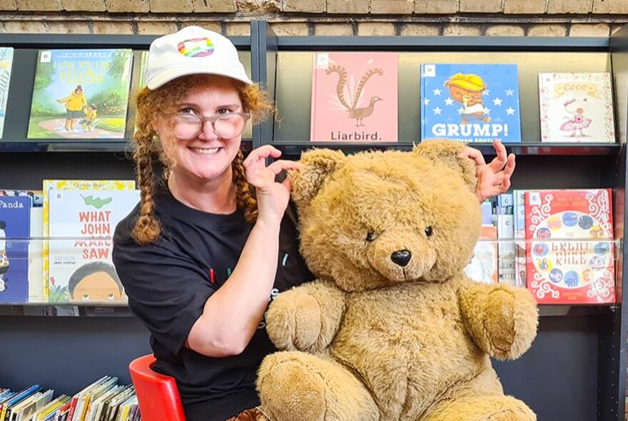 A smiling woman with long red plaits and a white cap in a library setting with a large bear on her knee. 