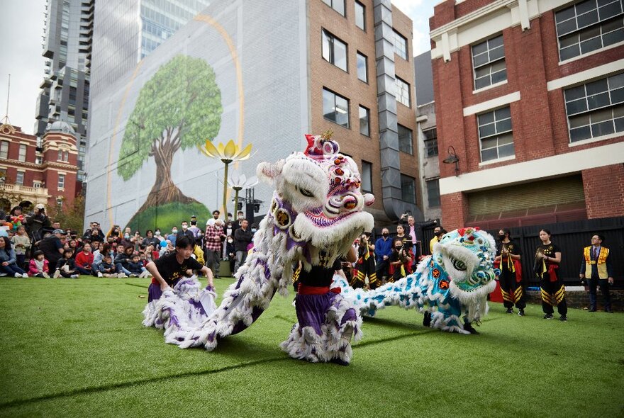 Lion dancers on an astroturf stage surrounded by people watching and city buildings.