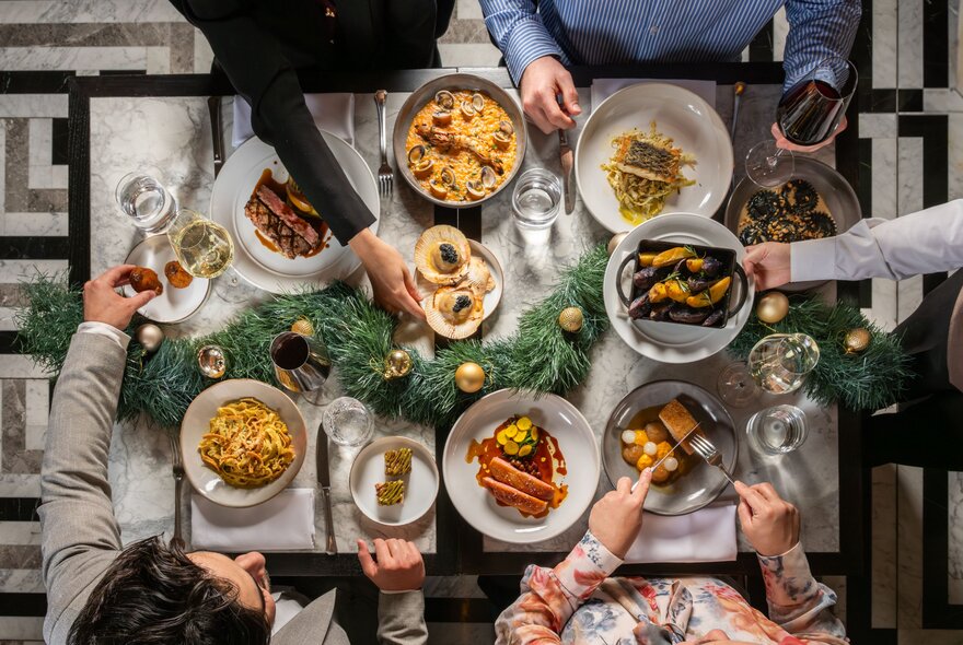 Overhead view of a table filled with plates of food and drinks, with people seated at it eating and drinking.