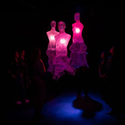 Three glowing, translucent pinkish-purple dress silhouettes with illuminated hearts float in a dark exhibition room space while people look on.
