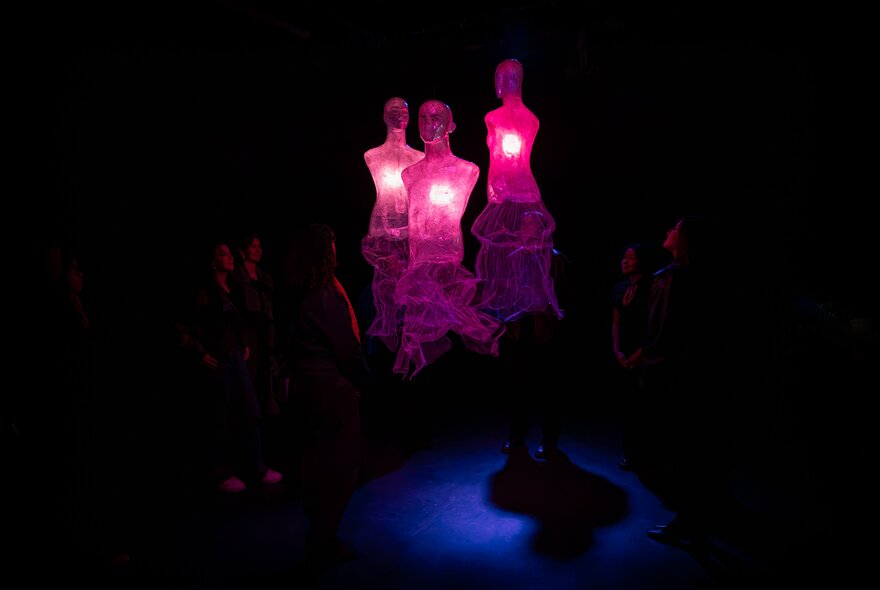 Three glowing, translucent pinkish-purple dress silhouettes with illuminated hearts float in a dark exhibition room space while people look on.