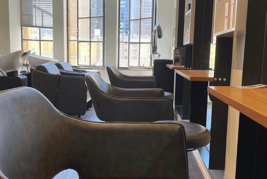 Interior of hair salon with club chairs, tables and mirrors and tall windows in the background.