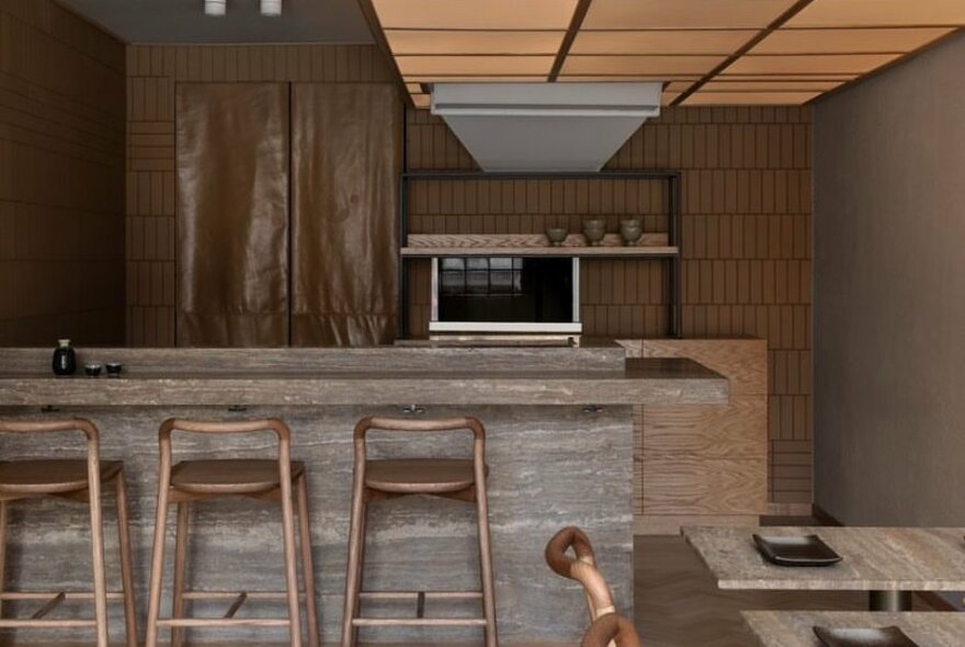 Modern and minimal interior of Waku Waku restaurant showing high stools at a kitchen counter, a small kitchen area behind, and dining tables to the side against a side wall. 