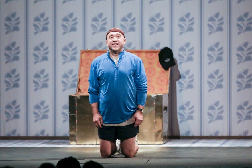 The actor George Kapiniaris kneeling on a theatre stage facing the audience, behind him is an open chest.