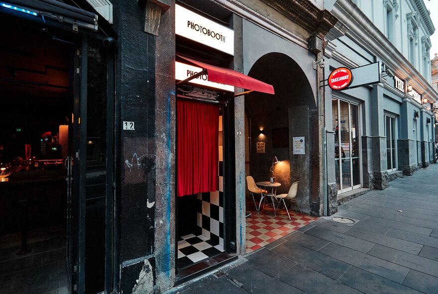 Vintage-style photo booth with red curtains and check floor, next to restaurant interior with similar design and arched windows.