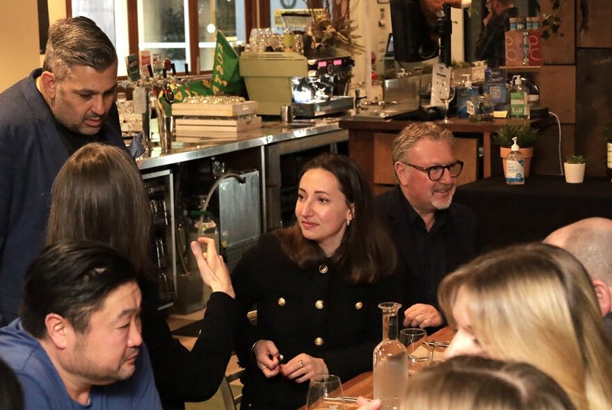 Dining patrons seated around a long table in a restaurant, conversing with each other and talking to one of the waiting staff who stands behind the table.