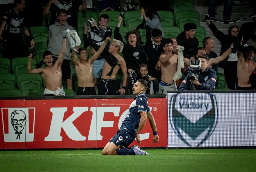 A soccer player falls to his knees in front of a crowd of men, some with no shirts on. 