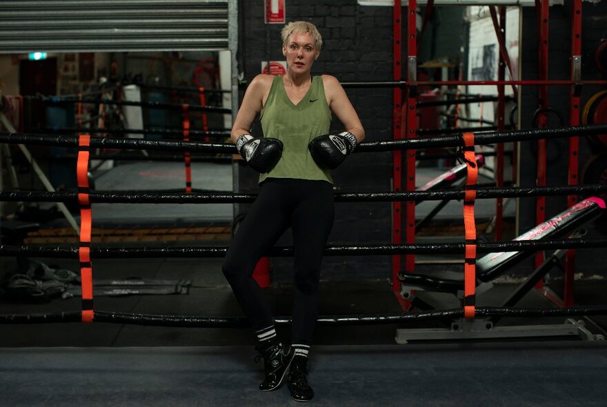 A woman wearing boxing gloves and a green singlet leaning back on the ropes of a boxing ring inside a boxing gym.