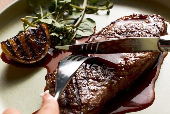 A knife and fork cutting into a steak on a dinner plate.