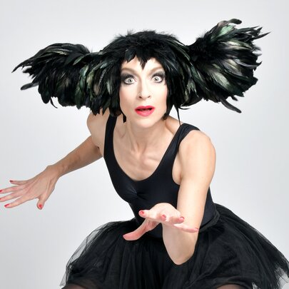 Performer dressed in a black tutu with winged hair, arms outstretched like a cat.