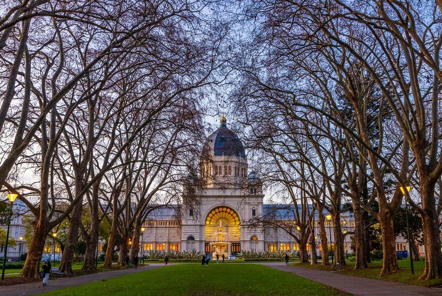 A grand domed building with rows of bare trees and a green lawn in front.