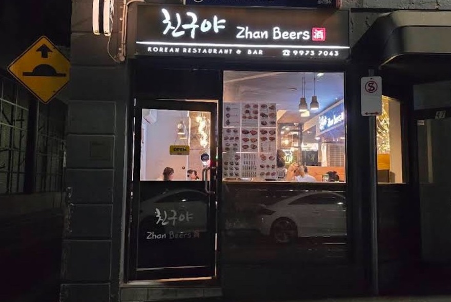 Exterior of Zhan Beers restaurant from the street at night, showing glass window and door, with a view into the lit interior.
